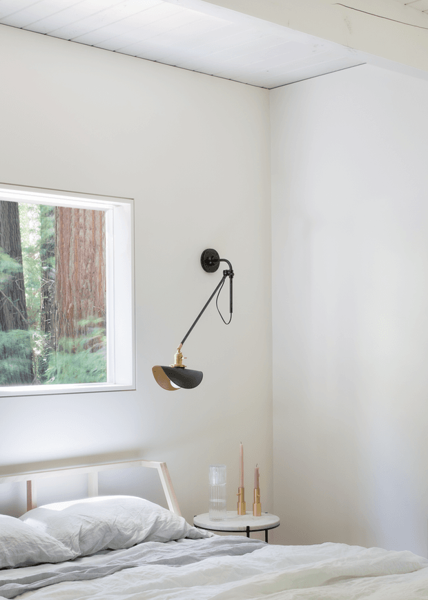A bright, minimal bedroom corner with a low white bed, rumpled white and pale grey bedding, a small round bedside table, and a wall-mounted articulated lamp. White walls and a white paneled ceiling create a clean, airy setting. A large window beside the bed looks onto dense redwood forest, while simple candles and glassware provide understated bedside styling.
