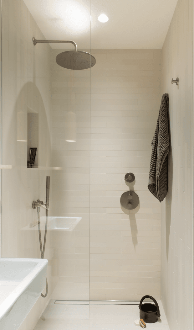 A compact, minimalist bathroom shower enclosure viewed through a frameless glass screen. The shower has white horizontal wall tiles, a white tiled floor, brushed metal rain shower and controls, a built-in wall niche, and a white freestanding or partially visible bathtub along the left edge. A dark textured towel hangs on the right wall, with a small black bucket and brush on the floor. Recessed ceiling lighting provides a clean, warm-neutral atmosphere.