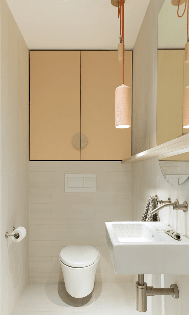 A compact contemporary bathroom viewed head-on, featuring a wall-mounted white ceramic basin with exposed chrome plumbing, a matching white toilet, white small-format tiled walls and floor, and warm beige overhead cabinetry with circular handles. A large mirror and slim shelf run along the right wall, while a suspended cylindrical pendant adds a warm decorative accent.