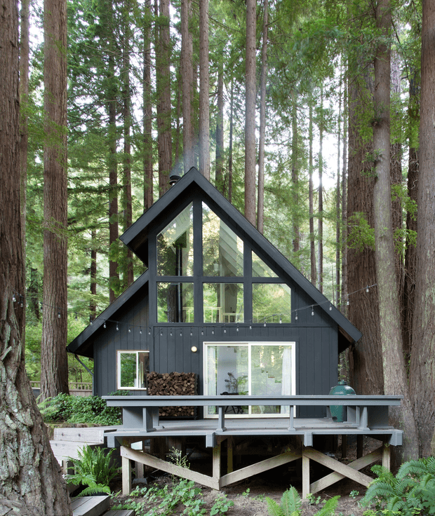 A compact black A-frame cabin set among tall redwood trees, with steep pitched rooflines, a dramatic glazed front gable, dark vertical exterior cladding, and a raised timber deck supported on timber bracing. String lights, stacked firewood, outdoor seating, and surrounding ferns create a secluded woodland retreat.