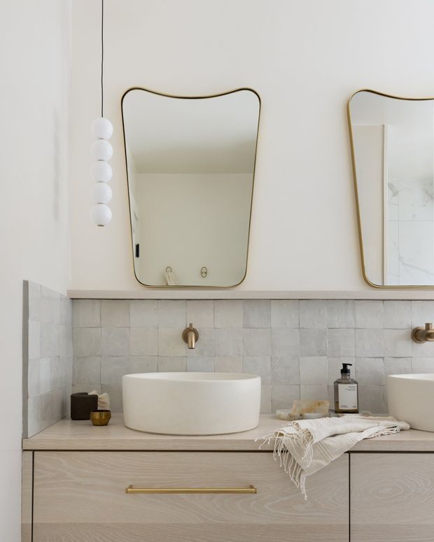 A refined contemporary bathroom vanity with a double-basin arrangement, pale timber cabinetry, a stone-look tiled splashback, wall-mounted brass tapware, and white vessel basins. Organic-edged mirrors with slim brass frames sit above the vanity, alongside a sculptural vertical globe light. The palette is soft and neutral with warm metallic accents and understated natural textures.