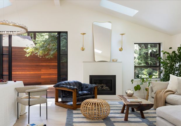Bright, refined living room with a cream fireplace surround, black-framed windows and glazed doors, a blue leather armchair, pale upholstered seating, a timber coffee table, a woven rattan ottoman, and a blue-and-cream rug. The room opens to a timber privacy screen and greenery outside, with skylights providing abundant natural light. Mid-century-influenced furniture and sculptural lighting complement the clean contemporary interior.