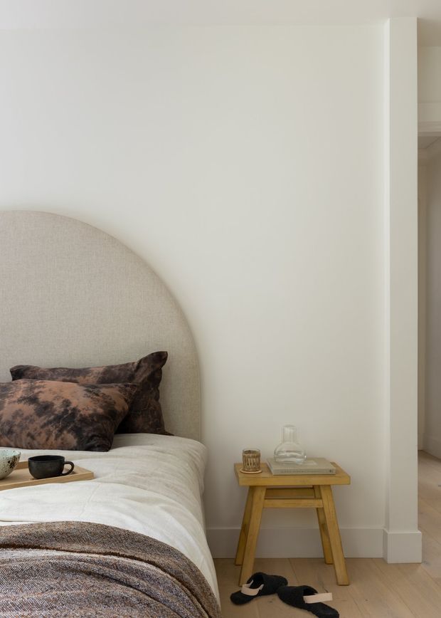 A softly styled bedroom corner with a curved upholstered headboard, neutral bedding, dark patterned pillows, and a small timber bedside table holding a glass carafe and cup. Pale walls, light timber flooring, and simple white trim create a calm, warm, minimalist setting. An adjacent hallway or room is visible through the opening at right.