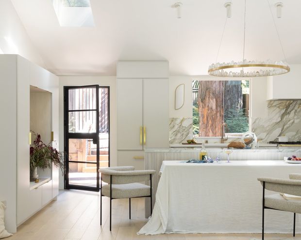 Bright, refined kitchen-dining space with a large central island draped in white fabric, pale cabinetry, marble-look splashback, light timber flooring, and a sculptural circular pendant light. Black-framed glazed doors open to an outdoor area, while a skylight and large window bring in abundant natural light. The restrained palette and curved upholstered stools create a soft contemporary interior.