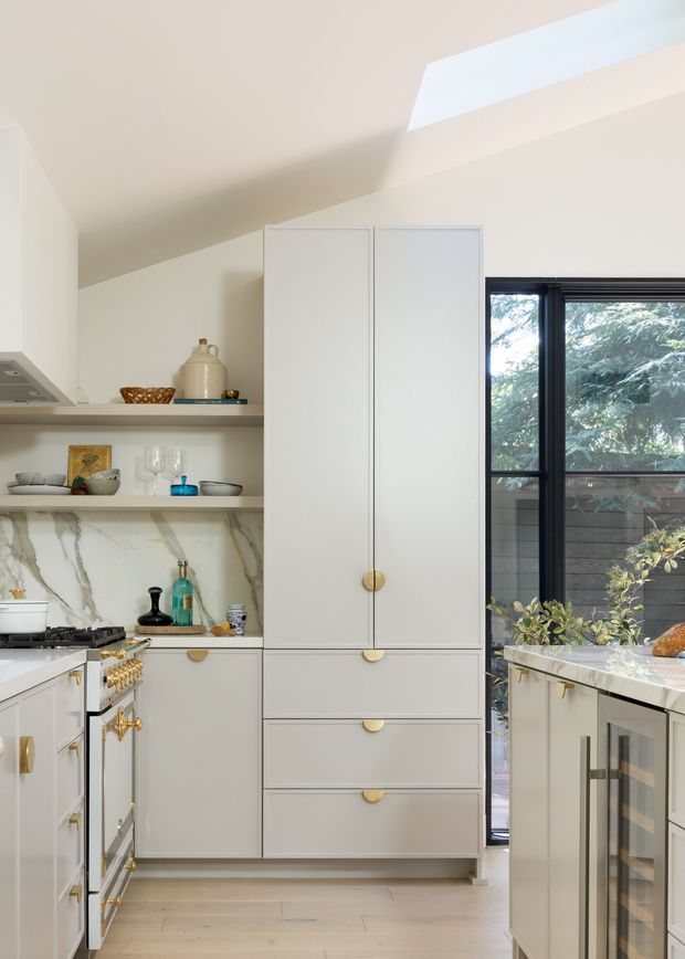 Bright, compact kitchen with a warm white and pale timber palette. The room features full-height white cabinetry with brass or gold knobs and pulls, open shelving displaying ceramics and glassware, a marble-look splashback, pale timber flooring, a gas cooktop and rangehood, and a dark-framed window opening to greenery. A skylight brings additional natural light into the sloped ceiling.