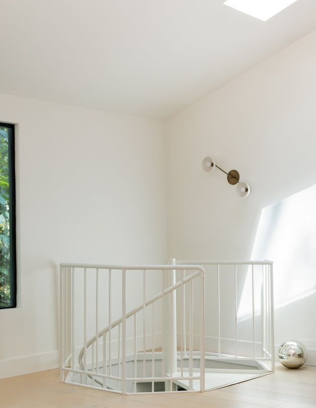 Bright minimalist stair landing with a white-painted spiral stair balustrade surrounding an open stairwell. The space has smooth white walls and ceiling, pale timber flooring, a slim dark-framed window with greenery outside, a ceiling skylight, and a sculptural wall light. Strong natural light creates an airy, clean atmosphere.