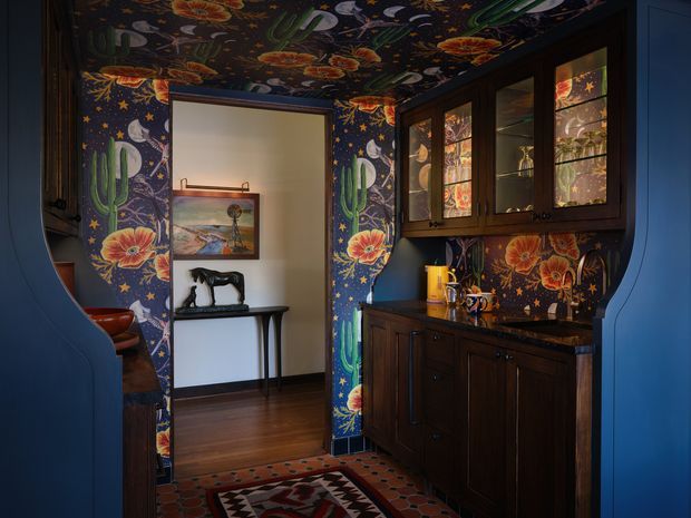 A richly decorated compact bar or beverage station framed by deep blue curved cabinetry. The walls and ceiling are covered in a vivid botanical wallpaper featuring cacti, flowers, stars, and birds. Dark wood lower cabinetry and glass-front upper cupboards sit beneath a dark stone or wood-look benchtop, with colourful glassware and accessories. The opening leads to a timber-floored hallway or adjoining room with a narrow console, artwork, and sculpture. The overall composition is eclectic, dramatic, and highly decorative.