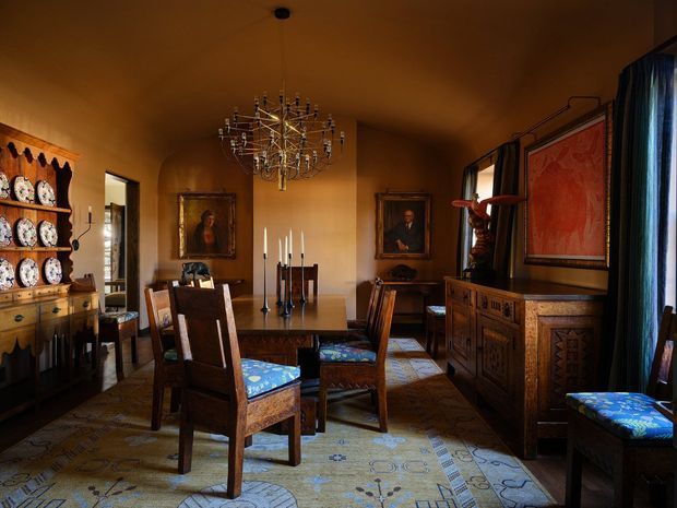 A richly furnished formal dining room with warm ochre walls, a large ornate chandelier, dark carved timber dining furniture, patterned upholstered chairs, antique-style cabinetry, framed artwork, and a decorative patterned rug. The room has a traditional, historic character with layered decor and low, warm lighting.