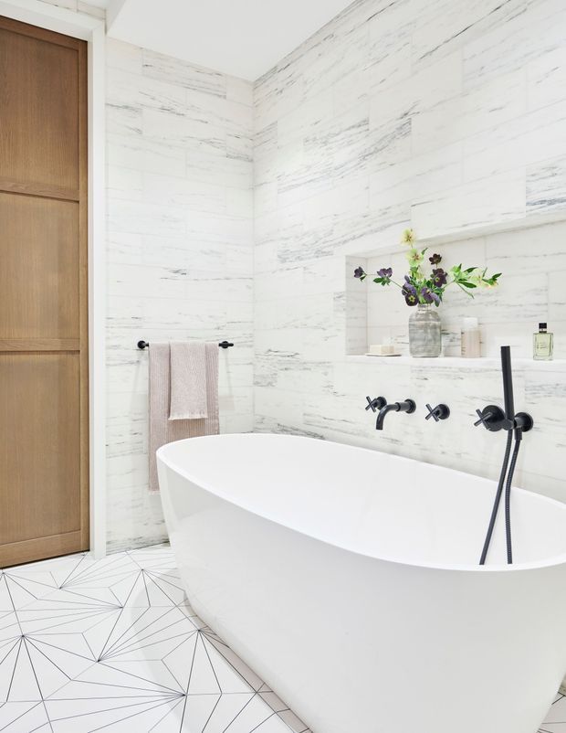 Bright contemporary bathroom featuring a freestanding white bathtub, white marble-look wall tiles with grey veining, a geometric white-and-grey floor tile pattern, matte black wall-mounted tapware and shower fittings, a recessed display niche, and a warm timber door.