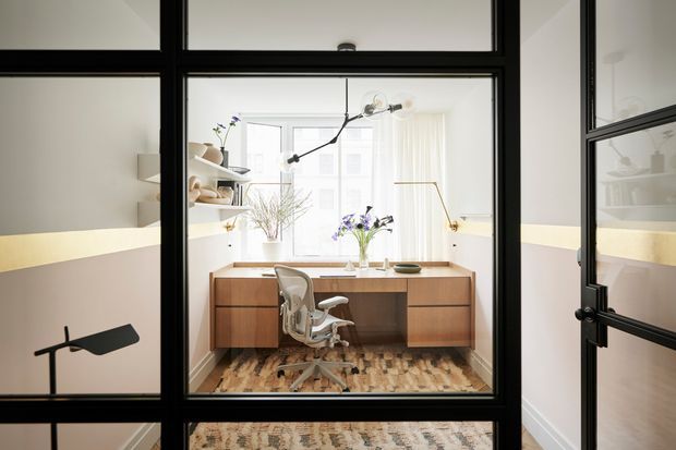 A bright, compact home office viewed through black-framed internal glass partitions and a glass door. The office contains a built-in timber desk with drawer cabinetry, a modern task chair, a sculptural pendant light, plants, and a large window with sheer curtains. Pale walls, warm timber flooring, and strong black framing create a refined contemporary workspace.