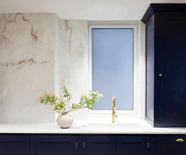 Close-up view of an elegant kitchen or wet-bar joinery installation, featuring dark navy-blue shaker-style cabinetry, a pale veined stone backsplash and benchtop, a brass gooseneck tap, and a narrow frosted or opaque window. The composition is bright and refined, with a small organic branch arrangement on the counter.