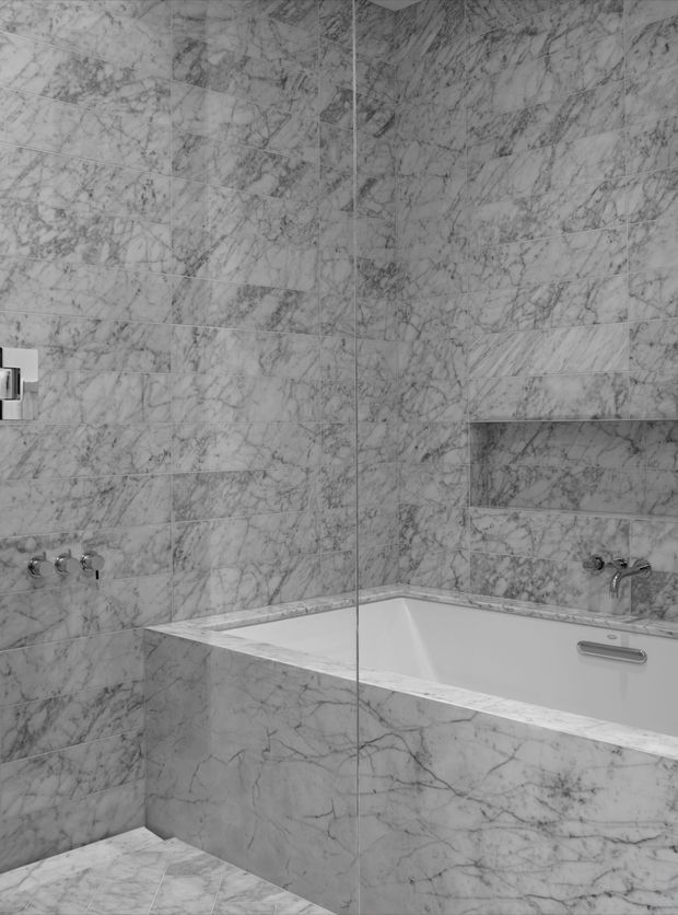 A monochromatic bathroom interior clad in large-format grey-white marble-look tiles with pronounced veining. A built-in white bathtub occupies the corner, surrounded by marble-faced walls and a matching tiled ledge. Wall-mounted chrome tapware and a glass shower panel are visible, creating a seamless, luxurious and highly cohesive finish.