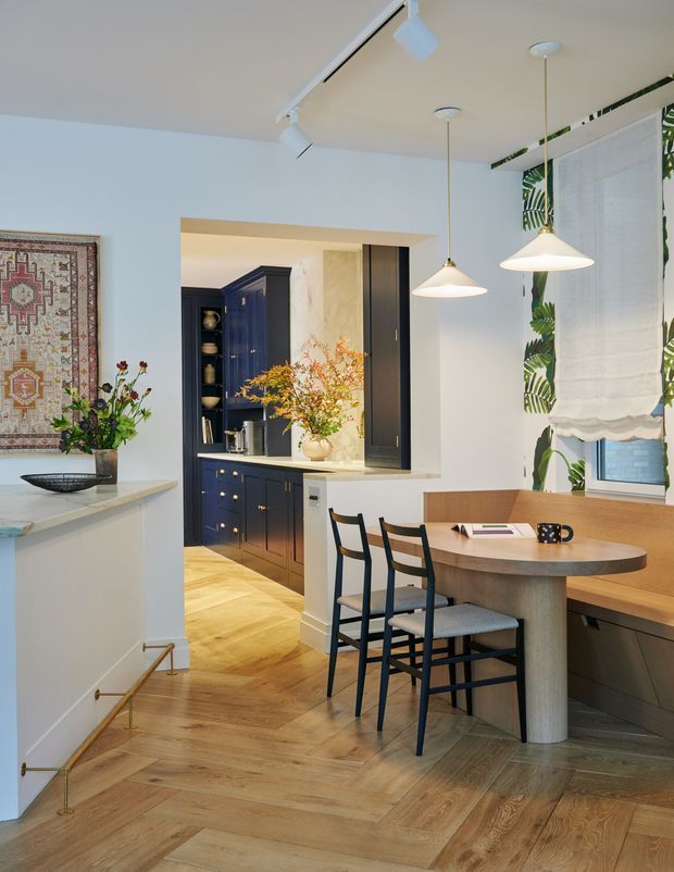 A bright, refined open-plan dining and kitchen interior. The foreground features a round timber dining table with black ladder-back chairs and an integrated timber bench beneath a large window. Beyond it is a compact kitchen with dark navy built-in cabinetry, brass hardware, pale stone benchtops, and open shelving. Light timber herringbone flooring, white walls, pendant lights, botanical-patterned window fabric, and framed textile artwork create a warm, layered interior.