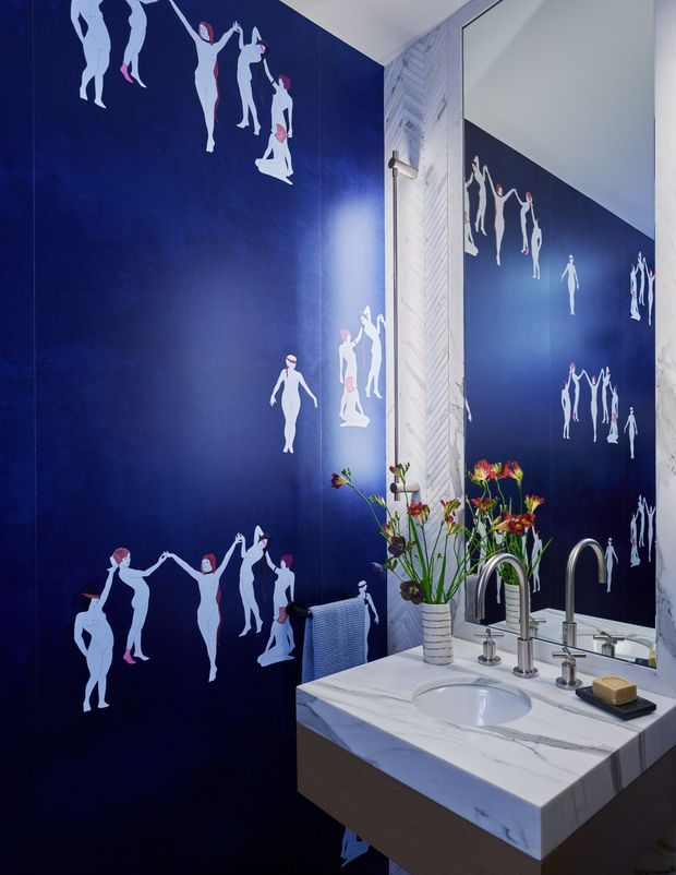 A compact, dramatically styled bathroom with deep navy walls covered in a playful white and pink dancing-figure mural. A large mirror reflects the artwork and vanity area. The room features a floating wood-toned vanity with a white marble benchtop and integrated basin, polished chrome wall-mounted and deck-mounted tapware, a small striped ceramic vase with flowers, and a white textured vertical feature panel beside the mirror.