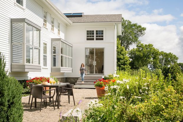 Bright exterior view of a white weatherboard residential building with projecting glazed window bays, a small gravel courtyard patio with outdoor dining furniture, steps leading to a glazed entrance, and abundant landscaped planting. The architecture has a simple traditional form with contemporary detailing and a low-pitched shingled roof.