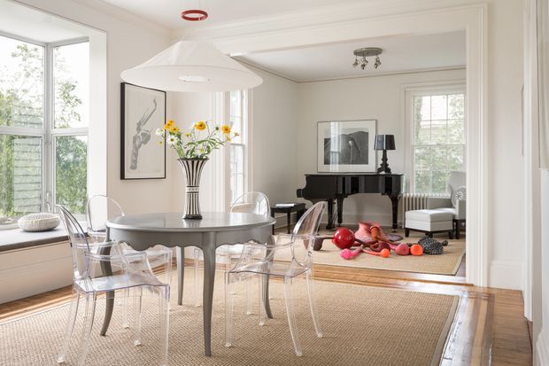 Bright, elegant open-plan dining and living space with a round glass-topped dining table surrounded by transparent acrylic chairs in the foreground. The room features large multi-pane windows, pale walls with traditional moulded trim, warm timber flooring, woven area rugs, a sculptural white pendant light, floral artwork, and a black grand piano in the adjoining sitting area. The overall aesthetic is refined and classic with a light, airy palette.