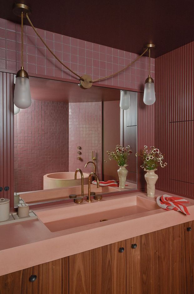 A richly coloured pink bathroom vanity with a wide integrated basin, pink square-tile walls, vertically fluted wall panelling, and a large mirror. The vanity has warm timber cabinetry with dark knobs, brass-toned taps and fittings, and sculptural pendant lights with frosted glass shades. Decorative vases with flowers and a folded red-and-white textile add accents to the retro, playful scheme.