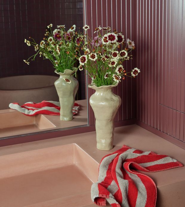A styled bathroom vanity scene with a large wall mirror, dusty rose or terracotta-toned tiled walls and integrated basin, and a cream ceramic vase filled with delicate white and burgundy flowers. A red-and-grey striped towel rests on the vanity. The image has a rich, warm, editorial feel with strong burgundy, blush, cream and red tones.