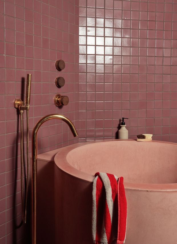 Close-up view of a pink tiled bathroom featuring a large freestanding or built-in rounded bathtub, rose-gold wall-mounted tapware and handheld shower, coordinated pink surfaces, and a red-and-white striped towel. The bathroom has a bold monochromatic palette with glossy square wall tiles and warm metallic fixtures.