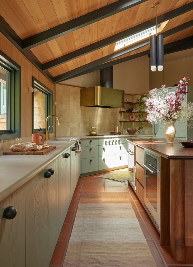 Warm, characterful galley kitchen with pale green lower cabinetry, dark round knobs, a wood-topped island and integrated appliances. The kitchen features a cream square-tile splashback, brass rangehood, brass tapware, terracotta floor tiles, timber-lined vaulted ceiling with exposed dark beams, and abundant natural light from skylights and windows. Open shelving, a large floral arrangement, and a runner add a lived-in, refined feel.