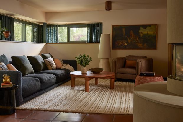 Warm, inviting mid-century-inspired living room with a large dark upholstered sectional sofa, leather armchair, round wooden coffee table, textured cream area rug, indoor plants, artwork, and expansive horizontal windows. A curved fireplace surround is visible at right, with terracotta-toned floor tiles and muted earthy finishes throughout.