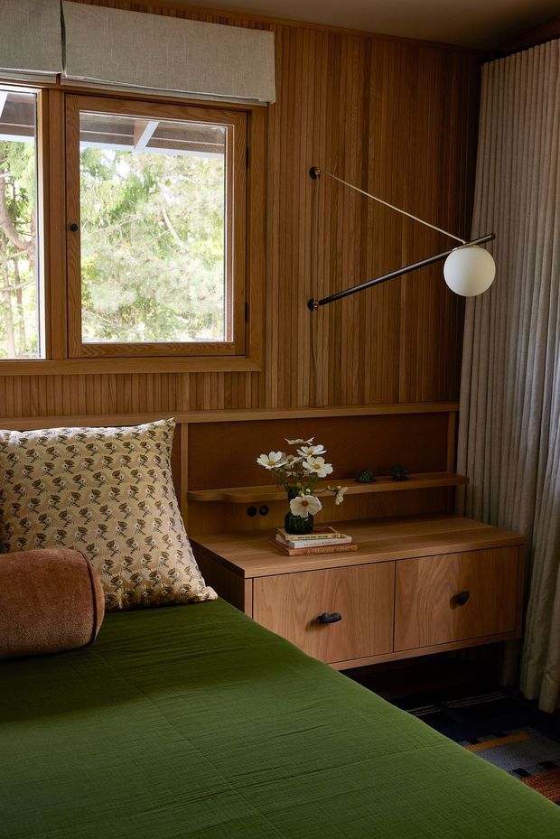 A compact, warmly styled bedroom corner with rich vertical timber wall panelling, a timber-framed window, and a green upholstered bed. A built-in timber cabinet and open shelf sit beneath a minimalist wall-mounted swing-arm lamp, with patterned cushions, books, flowers, and curtains adding character. The room has a distinctly mid-century feel and overlooks leafy greenery.