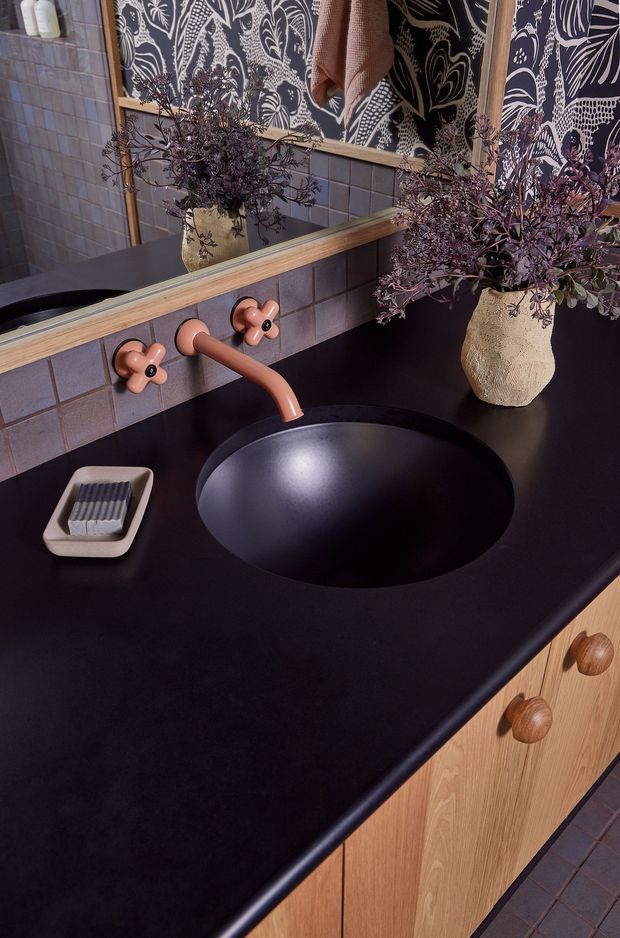 Close-up of a boldly styled bathroom vanity with a matte black oval vessel basin set into a dark countertop, terracotta-pink wall-mounted tapware, and light timber cabinetry with round wooden knobs. Muted blue-grey square wall tiles are paired with a large botanical patterned mirror or wall covering, a timber-framed mirror, and a decorative textured vase with foliage. The scheme is richly contrasted, graphic, and warm.