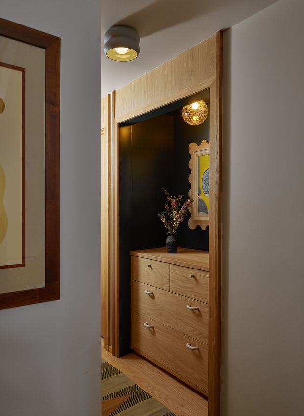 A narrow, warmly lit interior passage featuring a built-in timber dresser recessed into a dark feature wall. The custom joinery has multiple drawers, a timber surround and flooring, with a decorative pendant light, framed artwork, vase and flowers adding character. A partial framed artwork is visible on the adjacent wall.