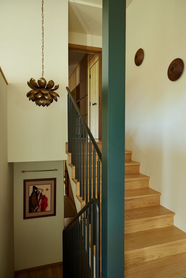 A tall, warm-toned stairwell viewed from an intermediate landing. Broad natural timber stairs rise along the right side, with dark metal balustrades and a prominent deep teal structural column. Cream walls are decorated with small circular wood wall pieces and framed artwork, while a sculptural bronze pendant hangs in the stair void. Timber panelled doors and joinery are visible at the upper landing.