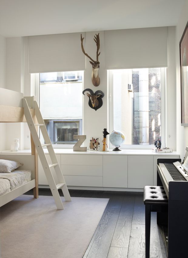 Bright, compact children's bedroom with a white bunk bed and ladder on the left, a long built-in white drawer unit beneath large windows, and a black digital piano with bench on the right. The room includes a pale area rug over dark timber flooring, roller blinds, a globe, toys and books, framed artwork, and decorative animal horns mounted between the windows. The overall design is clean, contemporary, and predominantly white with playful educational details.
