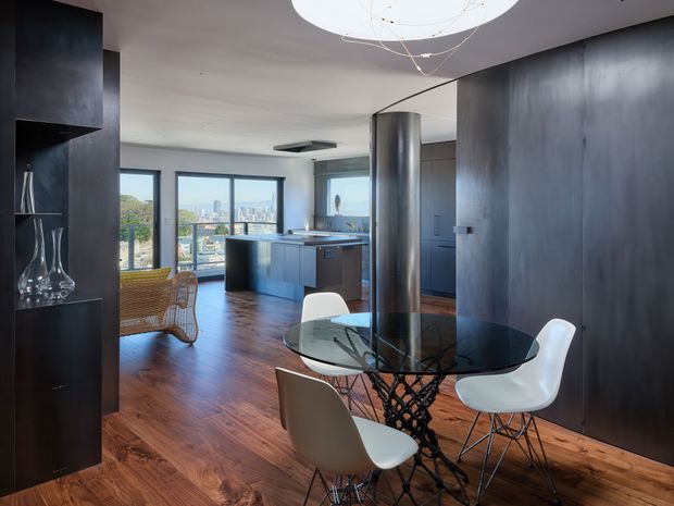 Open-plan contemporary apartment interior with a dining area in the foreground and kitchen beyond. The space features dark metallic-looking built-in cabinetry and wall panels, warm timber flooring, a glass dining table with sculptural black base, white and neutral modern dining chairs, a cylindrical metal-clad column, and large windows overlooking an urban skyline. A sculptural pendant light hangs above the dining table.