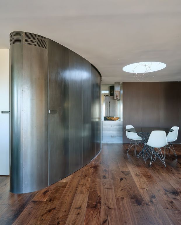 Interior view of a modern open-plan dining area with a striking curved brushed-metal partition or built-in wall at left. The room has rich timber flooring, dark wood wall cabinetry or panelling, a small kitchen zone in the background, and a glass dining table surrounded by white molded chairs. A circular ceiling opening or skylight with a sculptural light feature adds visual interest.
