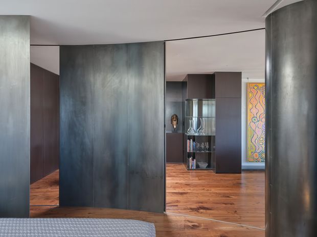 Interior view from a bedroom toward a dramatic open-plan living area, defined by large curved and flat dark metallic wall panels, dark built-in joinery, warm timber flooring, and a colourful framed artwork. The lower foreground shows the edge of a bed, while a glazed display cabinet and shelving are visible beyond.