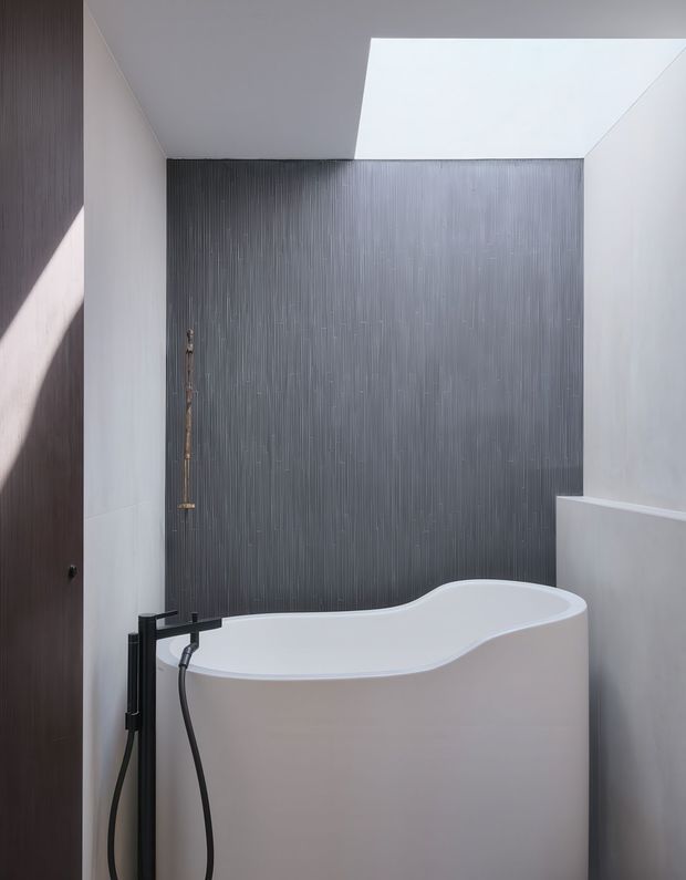 A compact contemporary bathroom featuring a freestanding sculptural white bathtub, matte black floor-mounted bath tap and handheld shower, dark vertically textured feature wall, pale smooth walls, and a large rectangular skylight. The space has a restrained monochromatic palette with strong natural illumination and clean geometric detailing.