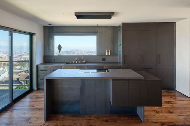Contemporary kitchen with a large central island, dark timber cabinetry, integrated appliances, and a stone benchtop. Full-height glazing and a wide window provide expansive city views, while warm timber flooring contrasts with the dark joinery. The room has a clean, refined, minimalist appearance with abundant natural light.