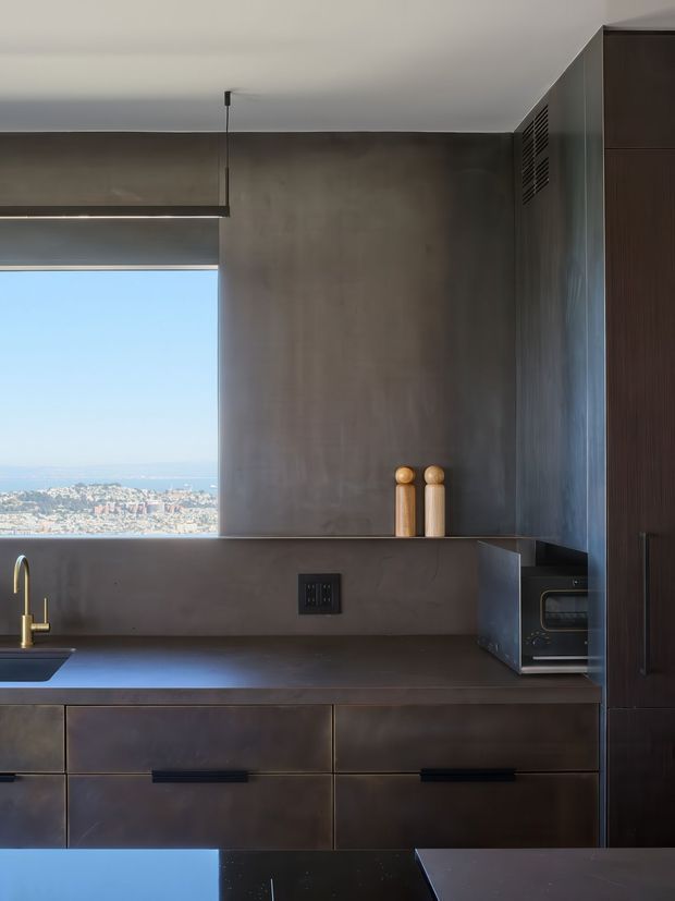 A moody contemporary kitchen with dark bronze-grey cabinetry and matching benchtops, a brushed or patinated metal-look backsplash and wall finish, integrated appliances, and a compact undermount sink with a brass-toned tap. A large horizontal window brings in natural light and overlooks a distant coastal urban landscape. Minimal pendant lighting and simple cylindrical decorative vessels reinforce the restrained, industrial-inspired aesthetic.
