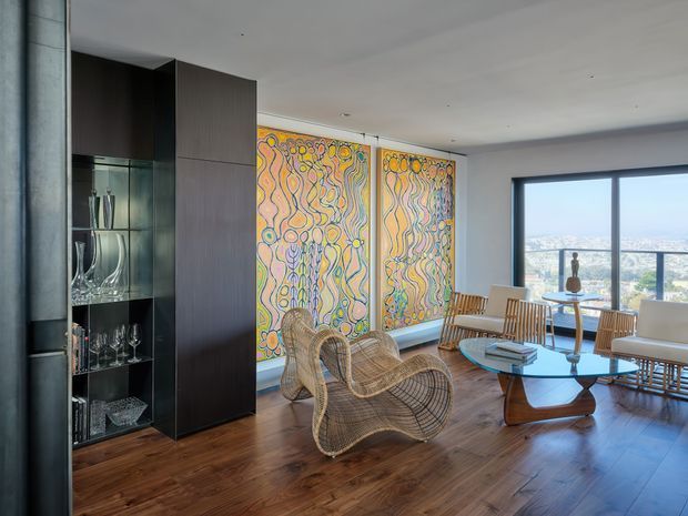 A bright contemporary living room with expansive city-view glazing, warm timber flooring, dark built-in display cabinetry, woven rattan lounge chairs, a low sculptural coffee table, and a light sofa. Two large colourful abstract artworks form a prominent feature wall, while a dining area sits near the window.