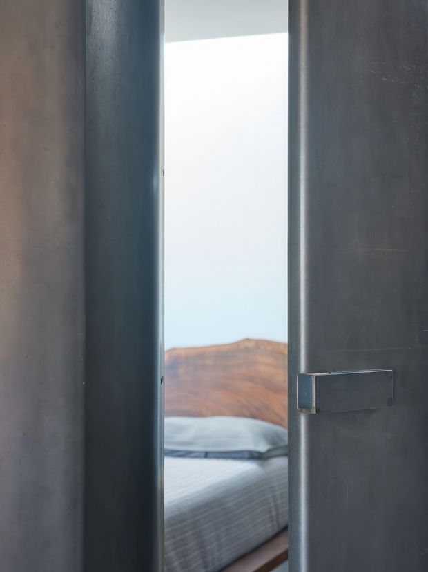 A narrow view through a partially open dark metal-framed door into a bedroom. The room contains a bed with grey striped bedding and a curved warm-toned wooden headboard, while the surrounding door and walls create a stark, monochromatic architectural frame.