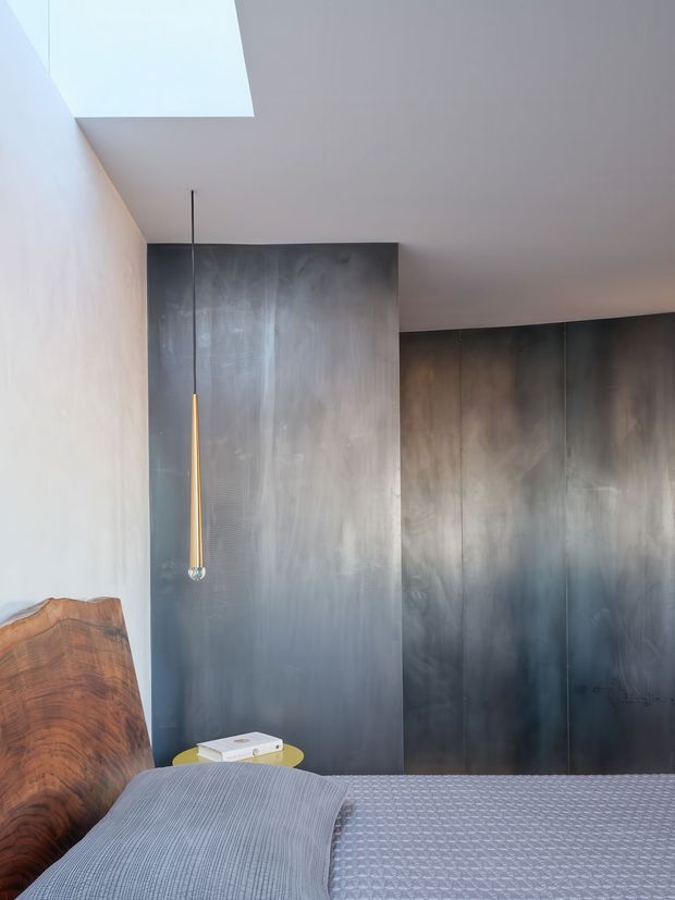 A moody modern bedroom corner with a low bed in the foreground, a sculptural warm-toned wood headboard or chair at left, dark smoky grey built-in wall panels, and a slender pendant light. A large skylight or roof opening brings soft daylight onto the pale plaster ceiling and wall. The restrained palette and minimal detailing create a sophisticated, atmospheric interior.