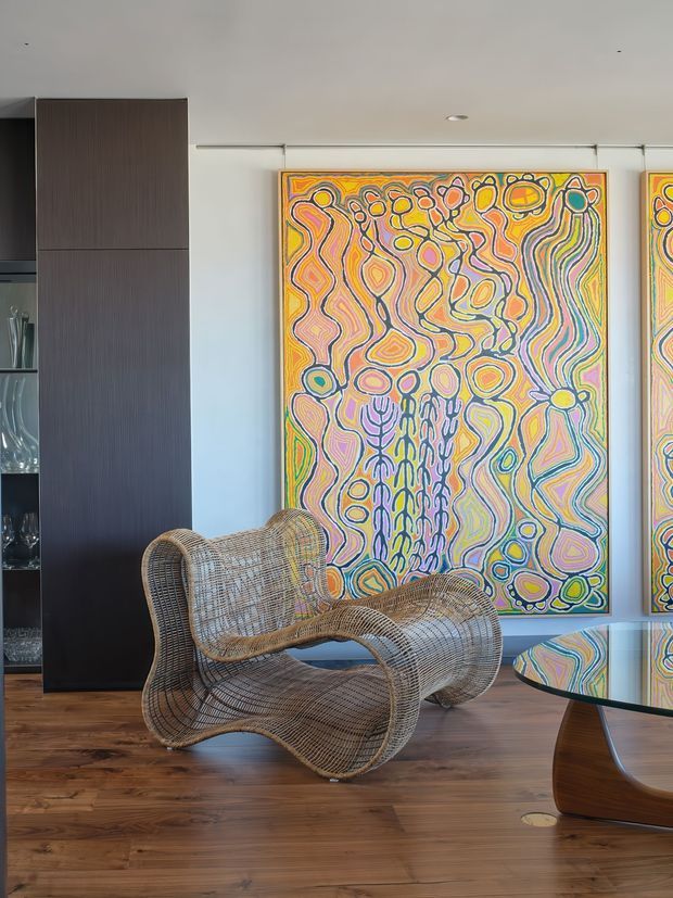 A styled contemporary interior corner featuring a sculptural woven lounge chair, a curved wood-and-glass coffee table, dark vertical-grain built-in cabinetry, and colourful abstract artwork mounted on a white wall. The room has warm timber flooring, recessed lighting, and a refined gallery-like atmosphere.