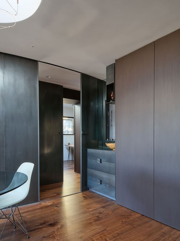 A sophisticated dark-toned interior hallway or dressing area with floor-to-ceiling ribbed wall panelling and concealed cabinetry. A black vanity with drawers and a vessel basin sits beside a full-height mirror, while a doorway opens toward a bright adjoining space with timber flooring and a small table. The room has a curved white ceiling feature, polished timber floorboards, dark joinery, and a partial view of a modern dining chair and table.