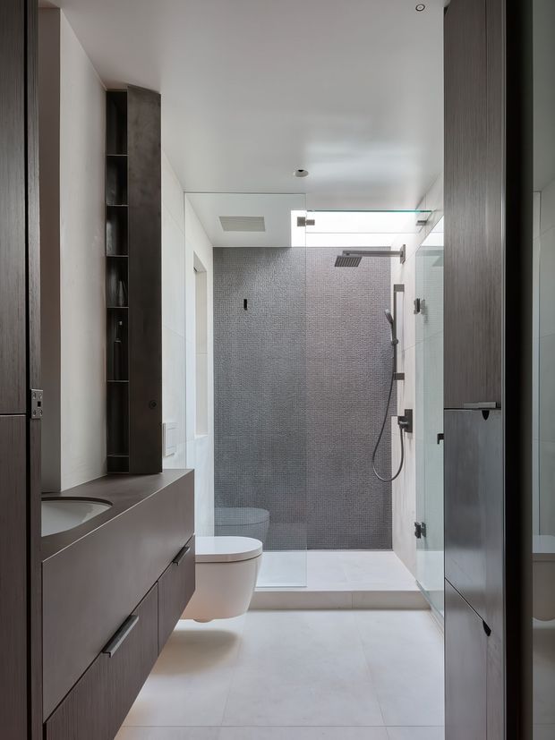 A narrow, contemporary bathroom viewed from the entry, featuring a floating dark timber-toned vanity with integrated basin, wall-hung toilet, glass-enclosed walk-in shower, large pale floor tiles, and a small-format grey mosaic tile shower wall. Tall dark vertical storage joinery and frameless glass panels reinforce the streamlined minimalist design.