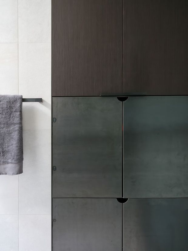 Close-up view of a minimalist bathroom wall with large pale tiles, a dark grey towel on a slim black towel rail, and floor-to-ceiling dark charcoal ribbed and smooth built-in cabinetry with concealed handles. The composition is highly linear and monochromatic, with a refined contemporary feel.