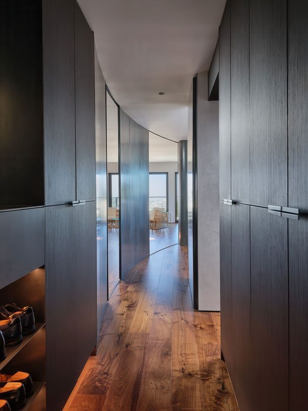 A narrow, sophisticated interior hallway lined with tall dark timber built-in cabinetry and concealed doors. The corridor has a warm natural timber floor, smooth light ceiling, subtle recessed lighting, and curved glass partitions leading toward a bright living area with large windows and ocean or city views. The design is highly streamlined and understated, with dark joinery contrasting against the warm flooring and daylight.