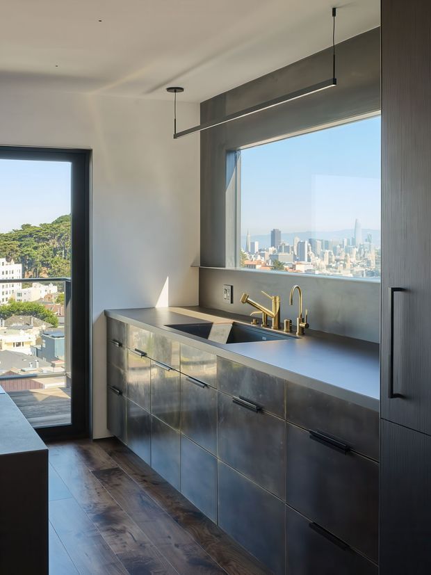 A compact, modern kitchen with a streamlined metallic built-in cabinet run, dark countertop, integrated sink, and brass-toned tapware. Large black-framed windows and a glazed door provide abundant daylight and expansive city views. A linear pendant light, dark timber flooring, and charcoal architectural surfaces create a refined urban look.