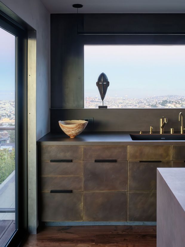 A compact, highly finished kitchen with dark bronze-toned fixed cabinetry, a dark benchtop, integrated sink and brass-toned tapware. Full-height glazing and a large picture window overlook a dense coastal cityscape. A sculptural bowl and decorative object sit on the counter and window ledge, while dark timber flooring and a pale stone island edge complete the composition. The space has a moody, refined contemporary character with strong natural light and expansive views.