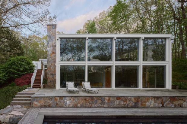 A modern glass-walled pavilion or house extension set within a wooded landscaped site. The facade features extensive floor-to-ceiling glazing in white frames, a low flat roof, and a substantial natural stone chimney and stone-clad terrace wall. Exterior steps lead up to a raised timber deck with outdoor lounge chairs, while a swimming pool is visible in the foreground.