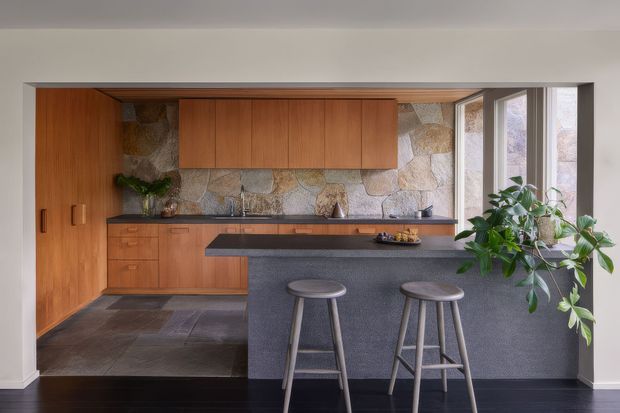 A warm, compact open-plan kitchen with extensive mid-tone timber cabinetry, a dark stone island with seating for two, and a natural irregular-stone splashback. Full-height glazing and a large indoor plant add light and organic character. Dark flooring contrasts with the warm joinery and pale walls.