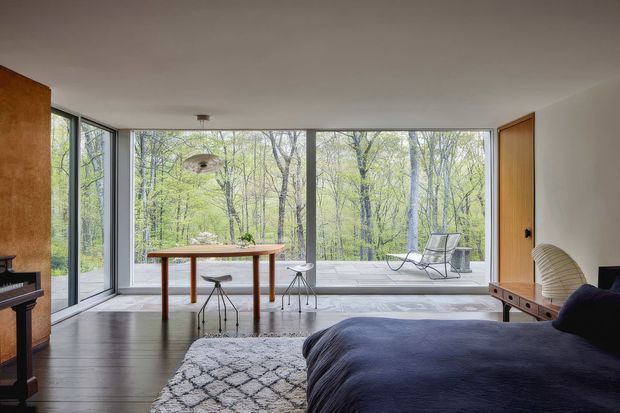 A serene, mid-century-inspired bedroom with a large bed in the foreground, a simple timber dining/work table and stools, dark timber flooring, and expansive floor-to-ceiling glazing overlooking a leafy wooded setting. The room includes warm timber wall and door panels, minimalist furnishings, a sculptural pendant light, and a lounge chair on the terrace beyond the windows.
