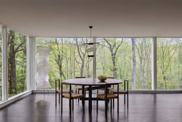A minimalist dining area with a dark wood or stained timber floor, a round dark dining table, and matching mid-century style chairs. Large floor-to-ceiling windows wrap around the room, providing expansive views into a leafy forest. A sculptural pendant light hangs above the table, while a tall white sculptural object stands near the glazing.